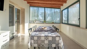 Un comedor con una mesa cubierta por un mantel estampado, vigas de techo de madera, ventanas amplias con cortinas y vista a árboles.