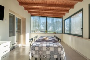 A dining area with a table covered in a patterned tablecloth, wooden ceiling beams, large windows with curtains, and a view of trees outside. - Villa 'Corbezzolo' with Shared Pool, Wi-Fi, and Air Conditioning (Galatone)