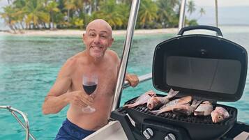 Un hombre asando pescado en un bote, con vista a una playa y palmeras al fondo.