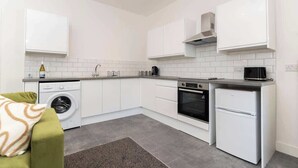 A compact kitchen with white cabinetry, a built-in oven, a washing machine, and a small refrigerator. - 4 Bedroom Apartment Central Redcar (Redcar and Cleveland)