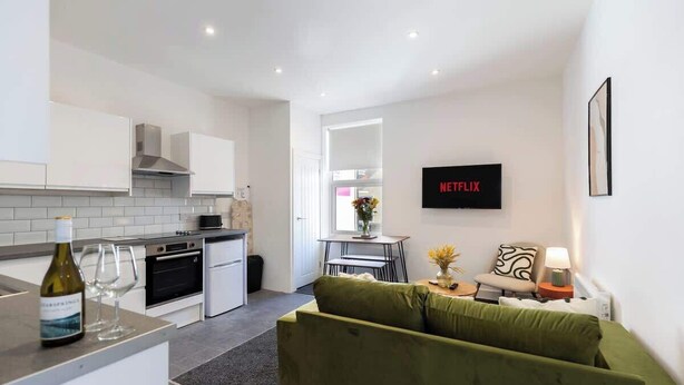 A modern kitchen with white cabinets, a dining area with a table and chairs, a green sofa, and a wall-mounted TV displaying 'NETFLIX'. - 4 Bedroom Apartment Central Redcar (Redcar and Cleveland)