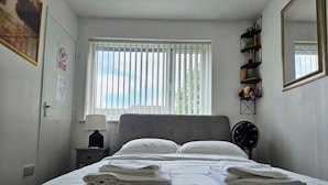 Een net opgemaakt bed met opgevouwen handdoeken, een nachtkastje met een lamp, een raam met zonwering en een wandspiegel.