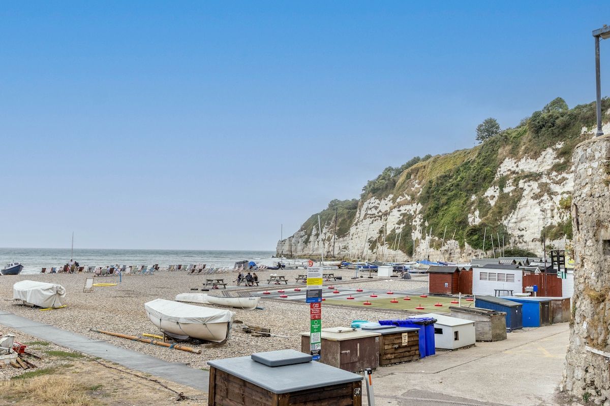 A pebbled beach with boats, a picnic area, and buildings on a cliffside.