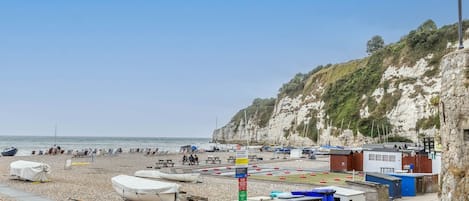 A pebbled beach with boats, a picnic area, and buildings on a cliffside.