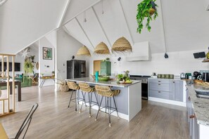 Eine moderne Küche mit einer zentralen Kochinsel, Barhocker, hängenden Körben und einem Essbereich, der durch eine Glaswand abgetrennt ist.
