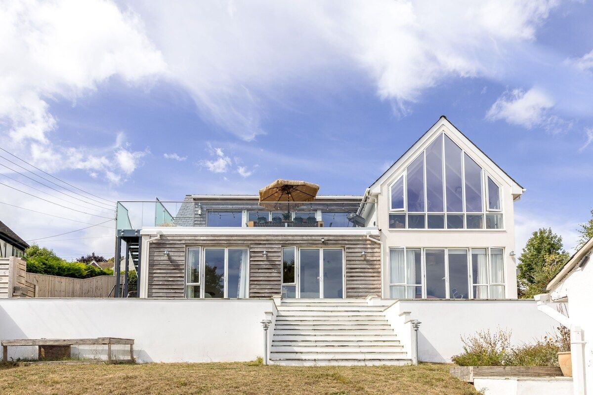 Una casa moderna de dos pisos con un amplio ventanal en el nivel superior, una terraza de madera y fachada blanca.