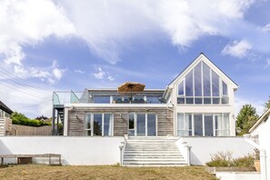 Ein modernes, zweistöckiges Haus mit großem Glasfenster im Obergeschoss, einer Holzterrasse und einer weißen Fassade.
