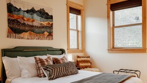 A bedroom with a bed, pillows, a blanket, a headboard, a wall tapestry, and windows. - Backroad Cabin • cozy, private retreat, fireplace, backyard with fire pit and string lights. (Emigrant)