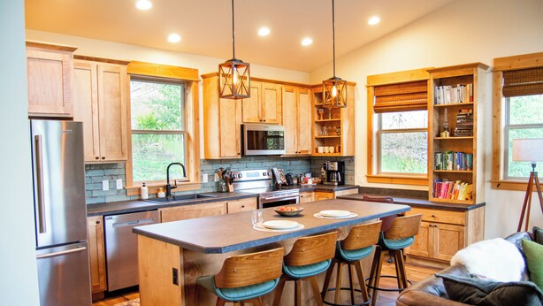 A modern kitchen with wooden cabinets, a central island with bar stools, stainless steel appliances, and pendant lighting. - Backroad Cabin • cozy, private retreat, fireplace, backyard with fire pit and string lights. (Emigrant)