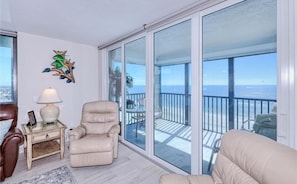A room with a beige leather chair, a glass table with a lamp, and a view of the ocean through large sliding glass doors.
