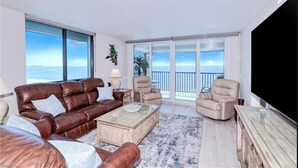 En rummelig stue med en brun lædersofa, beige lænestole og et glasbord. Store vinduer giver udsigt over havet.
