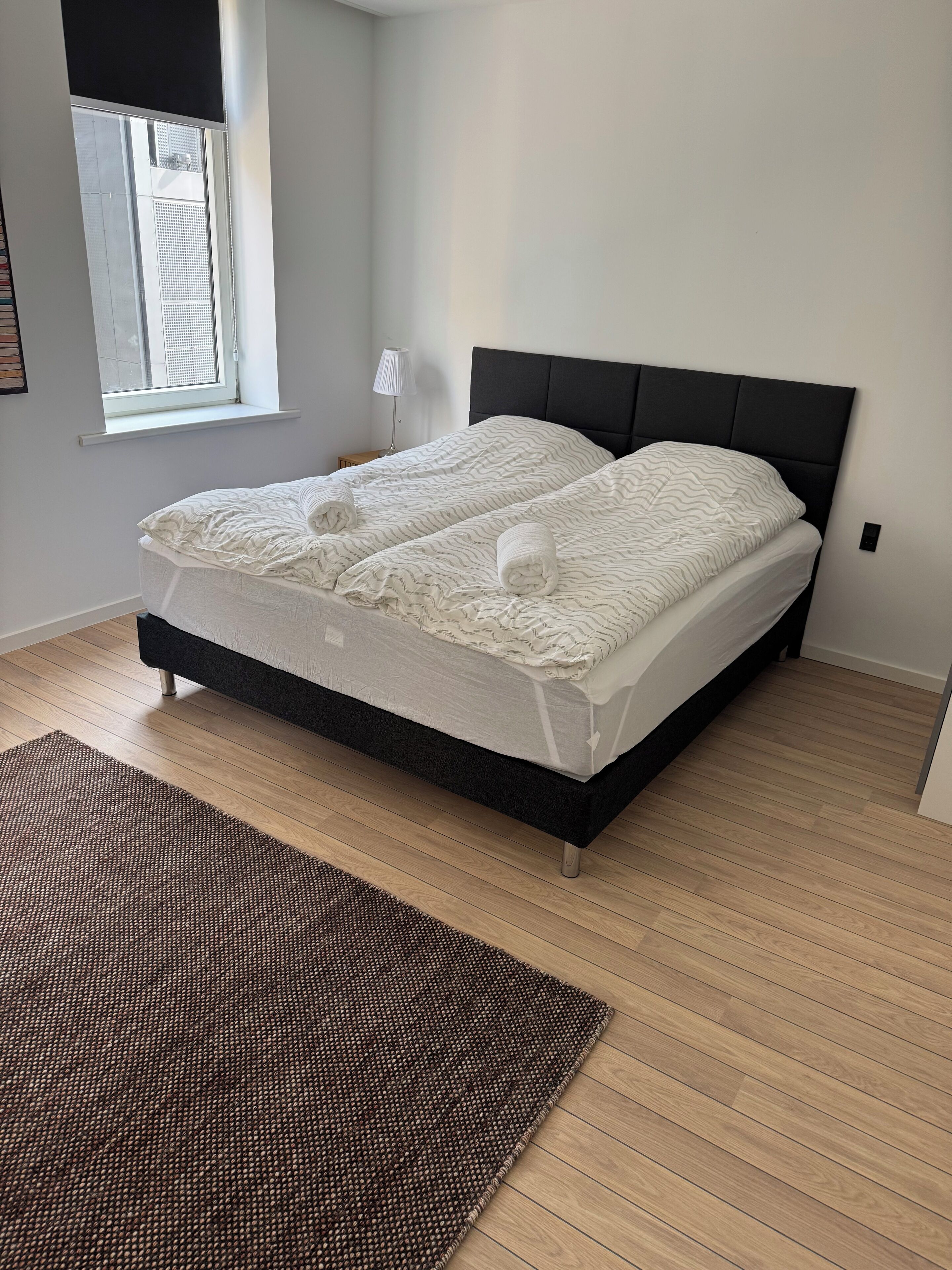A modern bedroom with a black headboard, two beds, a window, a lamp, and a brown rug.