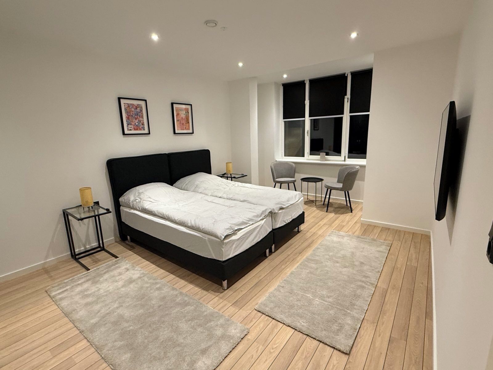 Un dormitorio moderno con una cama grande, dos mesitas de noche, un escritorio pequeño con una silla, una ventana con persianas y un televisor de pantalla plana montado en la pared.