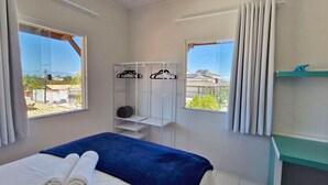 Una habitación de hotel con cama, un armario con perchas, una estantería y vistas a edificios y árboles.