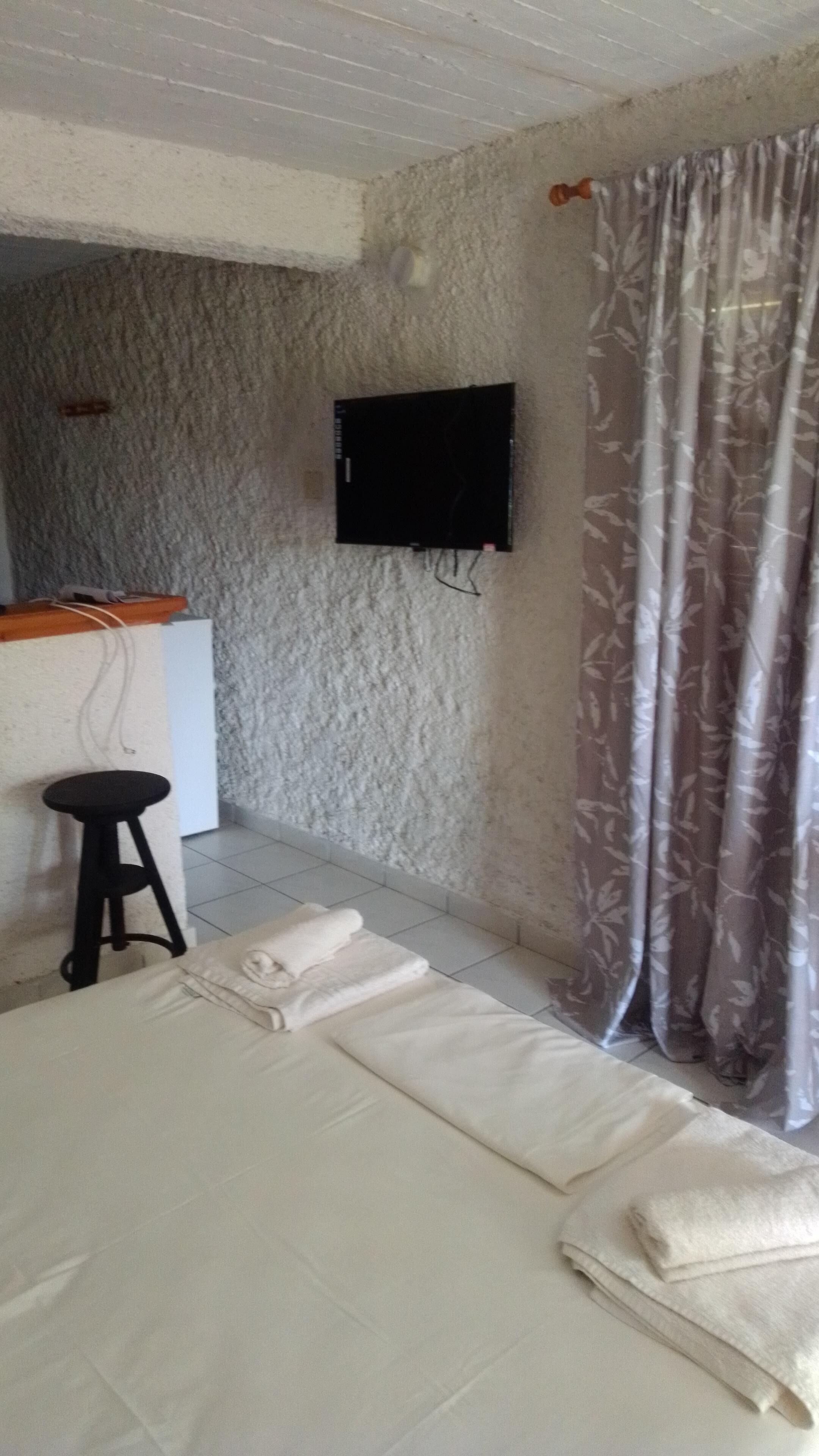 A room with a bed, a small stool, a flat-screen TV mounted on the wall, and a curtain with a leaf pattern.