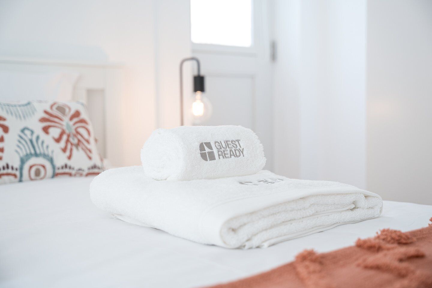 A neatly made bed with a folded white towel and a rolled towel with "GUEST READY" text, a patterned pillow, and a wall-mounted light fixture.