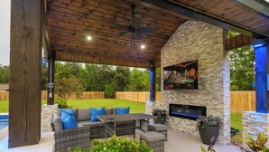 Un patio exterior techado con paredes de piedra, un televisor integrado y una piscina al fondo.