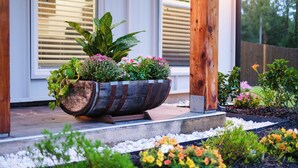 Una jardinera de madera con diversas plantas y flores sobre un borde de concreto frente a una casa.
