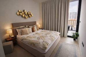A modern hotel room with a large bed, a headboard with a gold accent, a nightstand with a lamp, and a view of the city through a window.