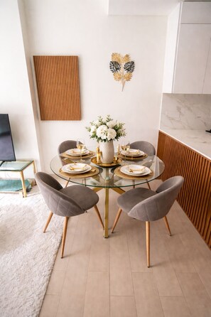 A dining area with a round glass table, grey upholstered chairs, a white vase with flowers, and a wooden accent wall.