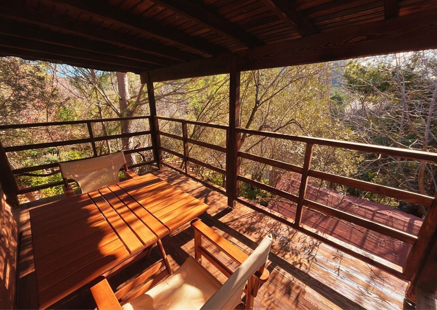 A wooden deck with a table and chairs, surrounded by a wooden railing, overlooking a forested area.