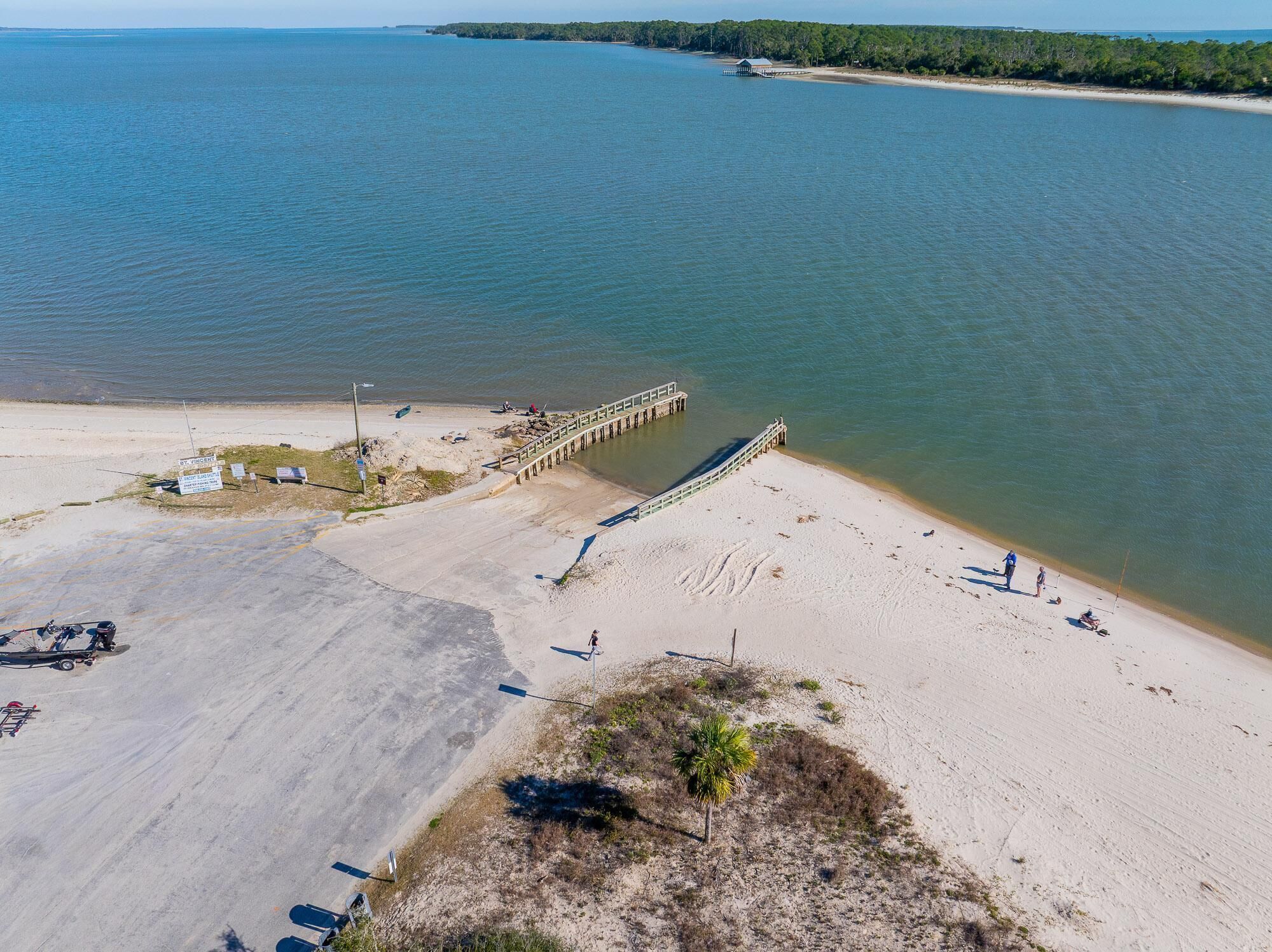 Ein Luftbild zeigt einen Strand mit einer hölzernen Pier, die sich ins Wasser erstreckt, einen Sandstrand und eine kleine Palme.