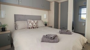 A bedroom with a bed, grey cabinets, a bedside table with a plant, and a window with blinds. - Meadows Park 4 (Hull)