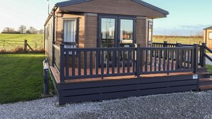 A single-story wooden cabin with a deck, large windows, and a fenced yard. - Meadows Park 4 (Hull)