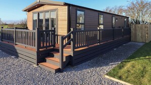A modern mobile home with a deck, large windows, and a gravel driveway.