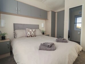 A bedroom with a bed, grey cabinets, a bedside table with a plant, and a window with blinds. - Meadows Park 5 (Hull)
