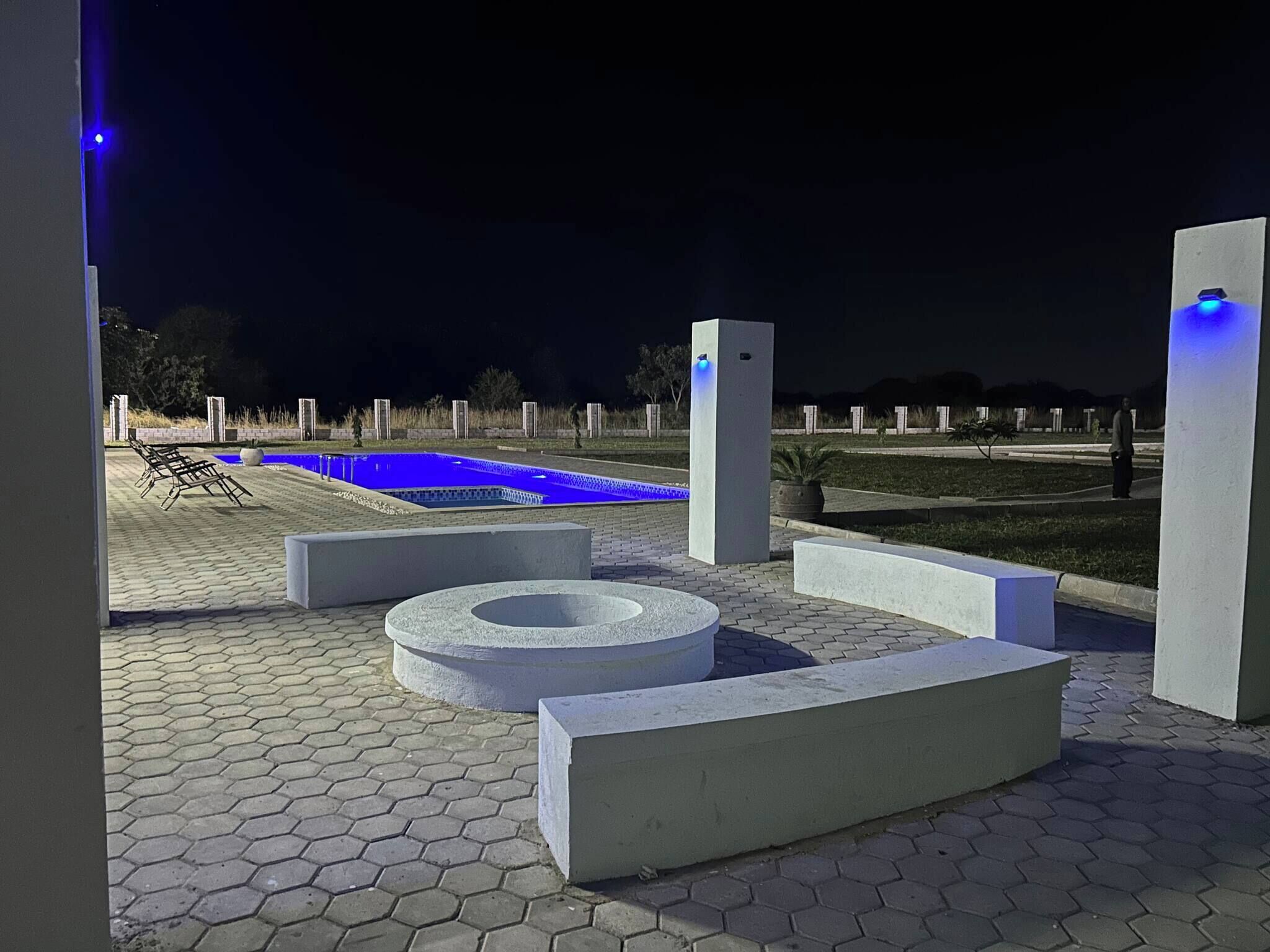 Un espacio al aire libre nocturno con piscina, sillones de descanso y estructuras de concreto geométricas.