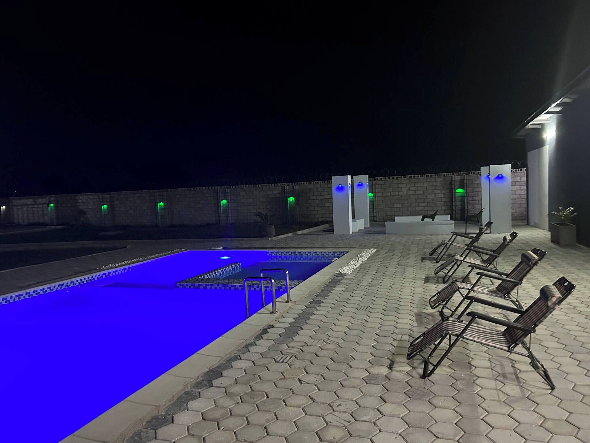 An outdoor swimming pool with hexagonal tiles, surrounded by lounge chairs and a brick wall with green lights.