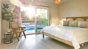 A bedroom with a large bed, a chair, a small table, a lamp, and a view of a swimming pool through a glass door. - @ Marbella Lane - Vibrant & Chic Designer Home (Fullerton)