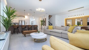 Ein modernes Wohnzimmer mit einer Couch, einem Couchtisch und Sesseln. Im Hintergrund ist eine Küchenzeile mit Schränken und einem Kühlschrank zu sehen.