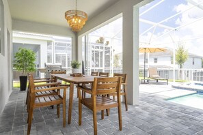 An outdoor patio with a wooden table and chairs, a hanging light fixture, and a view of a swimming pool and neighboring houses.