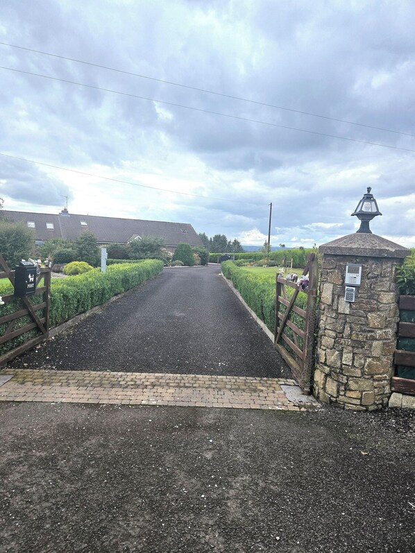 Une allée pavée mène à une entrée fermée par un portail, adossée à un mur de pierre sur lequel est posée une lanterne.