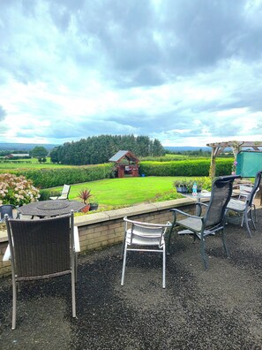 Une terrasse extérieure avec des chaises et une table en osier, donnant sur une pelouse bien entretenue et un bâtiment situé au loin.