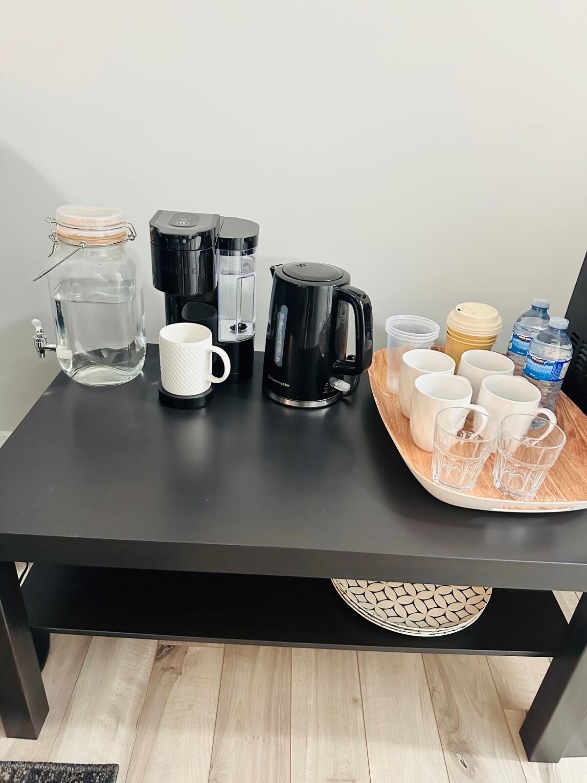 A black table with a water dispenser, a coffee maker, a mug, and a tray with cups and water bottles.