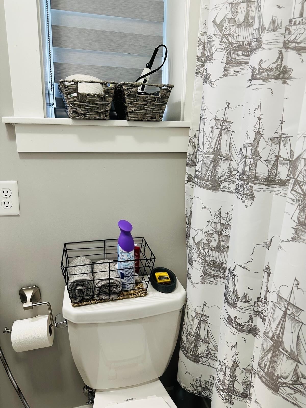 Ein Badezimmer mit Toilette, einem Fenster mit Korb, einer Duschvorhang mit nautischem Motiv und einem Toilettenpapierhalter.
