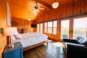 Une chambre dans une cabane en bois, avec un lit, une télévision, un ventilateur de plafond et de grandes fenêtres.