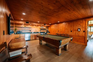 Une pièce aux murs lambrés, comprenant une table de billard, un coin bar et une cuisine.