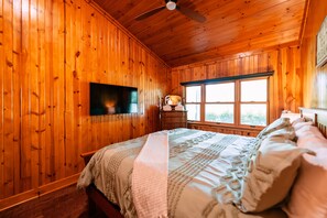 A wooden-paneled bedroom with a bed, a television, a window, and a ceiling fan.