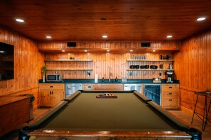 A pool table in a wooden-paneled room with a microwave, coffee maker, and shelves with glasses and bottles.