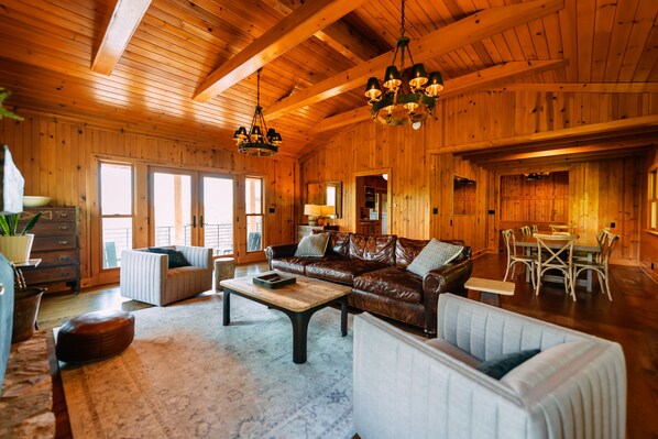 A spacious living room with a brown leather sofa, white armchairs, a coffee table, and a dining area with a table and chairs.