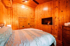 Une pièce aux murs lambrés, avec un lit, un téléviseur fixé au mur et une commode en bois.