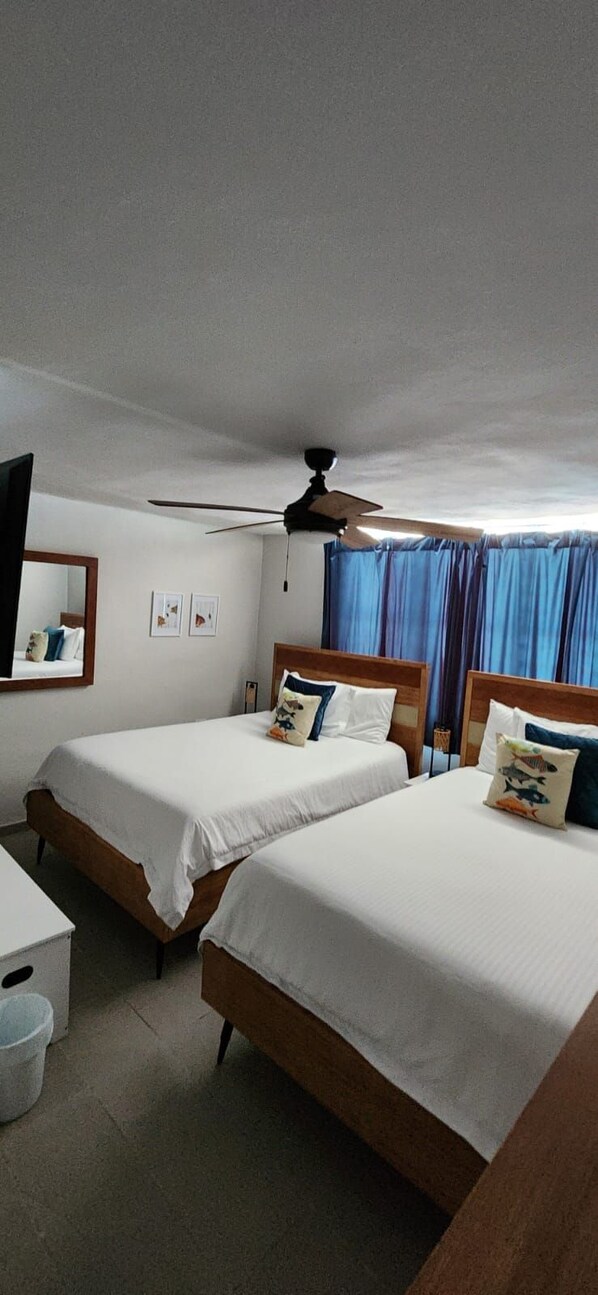 A hotel room with two single beds, a ceiling fan, a small table, and a window with curtains.