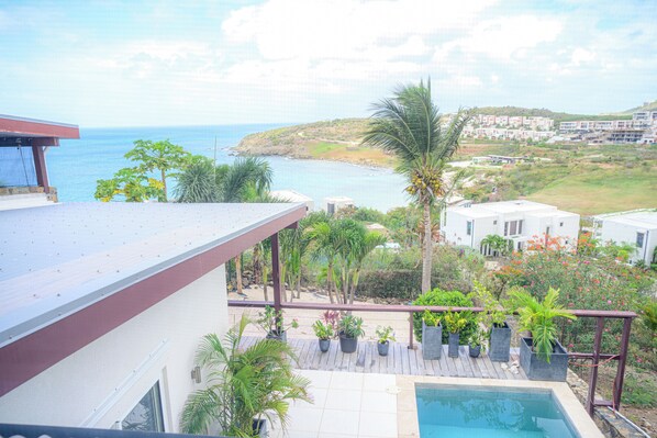 Zona de piscina con vista al mar, rodeada de plantas en macetas y una terraza de madera.