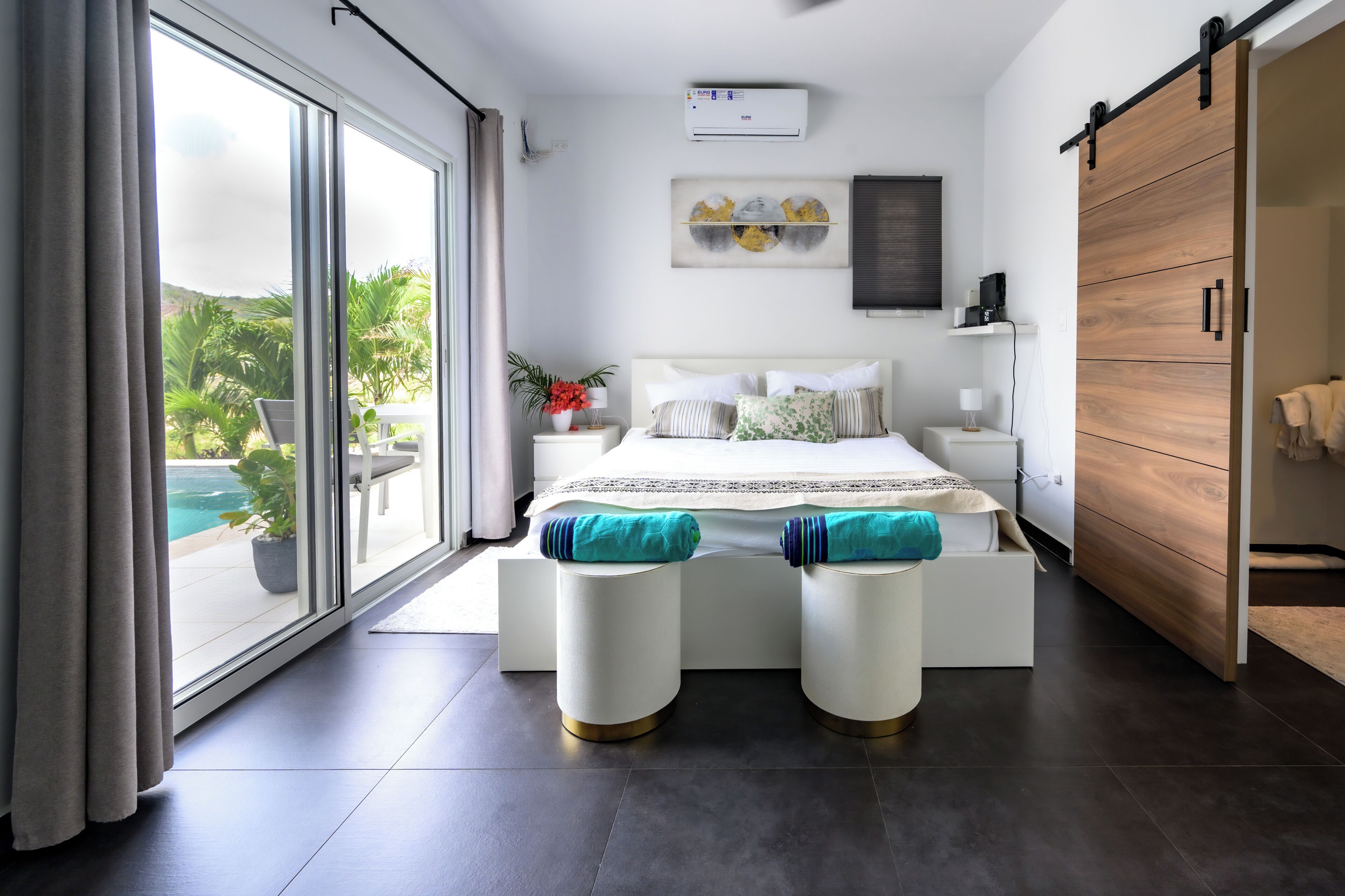 Une chambre moderne avec un grand lit, une armoire en bois à portes coulissantes, une table de chevet avec une lampe, et une vue sur une piscine à travers une porte vitrée.