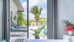 Una habitación de hotel con una cama, un balcón que ofrece vistas a una piscina y palmeras, y una planta en maceta sobre la mesita de noche.