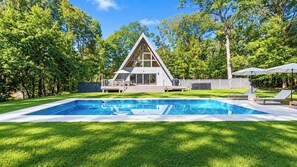 Una casa moderna con techo triangular, piscina y terraza con sillas de descanso.